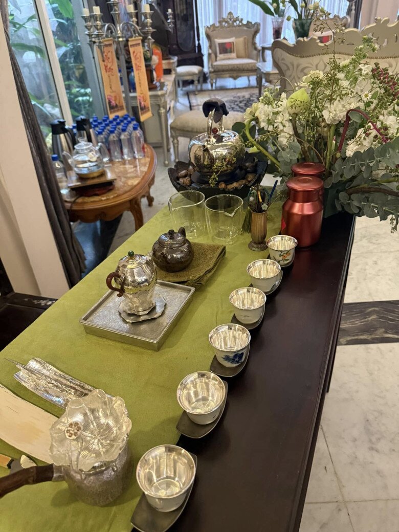 Bộ dụng cụ pha trà bằng bạc sáng bóng, gồm ấm, chén và khay trà, được bày biện ngay ngắn trên bàn phủ khăn xanh rêu. Phía sau là không gian phòng khách với bộ ghế cổ điển và những chi tiết trang trí cầu kỳ.