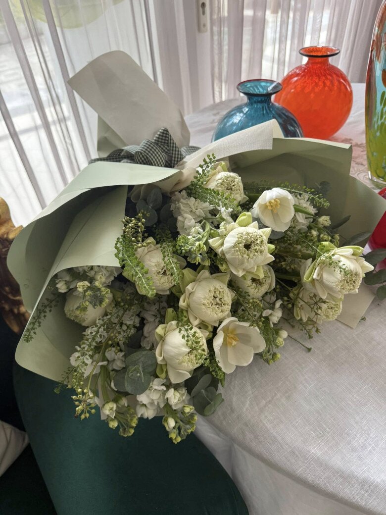 Một bó hoa trắng được bọc giấy màu xanh pastel, đặt trên bàn bên cạnh những bình thủy tinh màu sắc rực rỡ, tạo nên sự hài hòa giữa hiện đại và truyền thống.