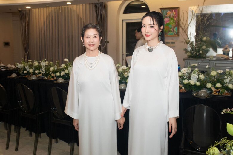 Giáng My và mẹ ruột diện áo dài trắng, tạo dáng trước bàn tiệc được trang trí đầy hoa tươi. Những bức tranh nghệ thuật treo trên tường giúp không gian trở nên sinh động và giàu cảm xúc hơn.