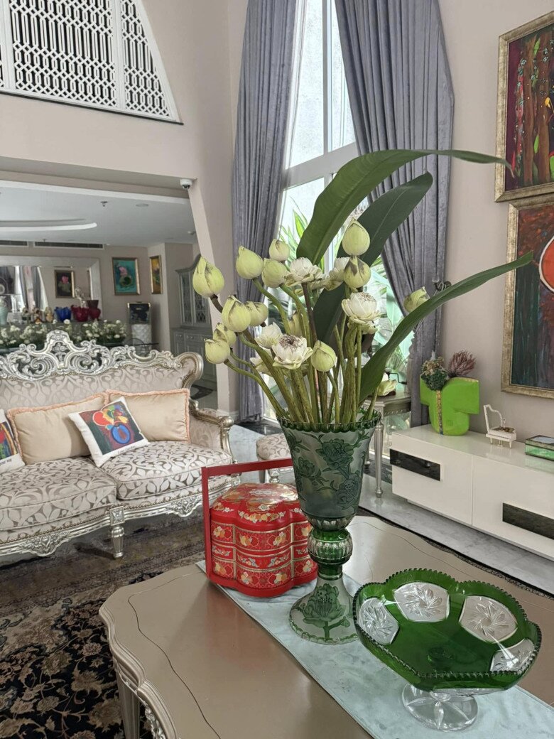 Khu vực phòng khách rộng rãi với sofa họa tiết bạc trang nhã, đi kèm những chiếc gối tựa nhiều màu sắc. Bình hoa sen trắng đặt trên bàn kính hợp tông với khay thủy tinh xanh lục mang phong cách hoài cổ.