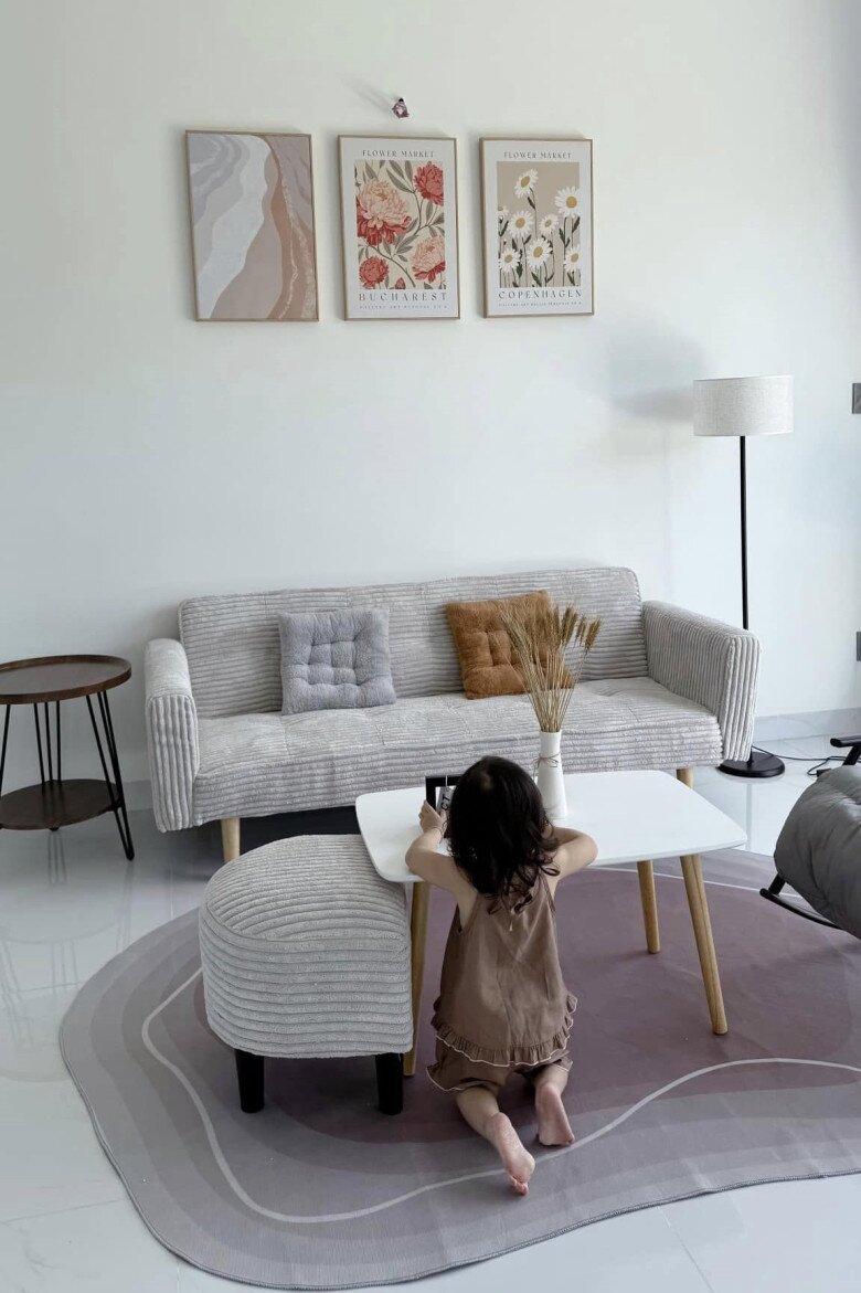 Bộ sofa màu xám nhạt kết hợp với bàn trà chân gỗ đơn giản. Trên tường là những bức tranh mang sắc thái mềm mại, hòa quyện cùng ánh sáng tự nhiên từ cửa sổ lớn.