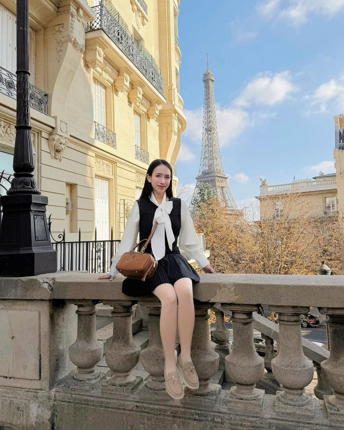 Con gái Minh Nhựa diện đồ phong cách preppy với váy yếm và sơ mi cổ nơ, kết hợp túi xách Miu Miu cùng giày loafer Lori Piana khi chụp hình ở Paris.