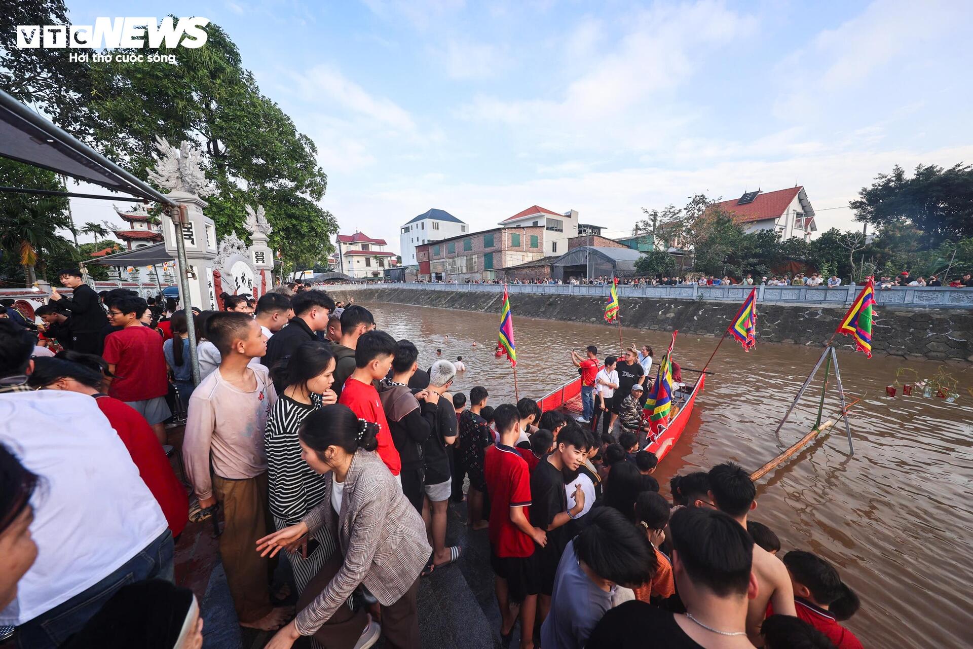 Một chiếc thuyền trang trí giấy đỏ được neo giữa dòng làm nơi điều hành trò chơi. Từ bờ, một cây cầu khỉ bằng tre vươn ra mặt nước.
