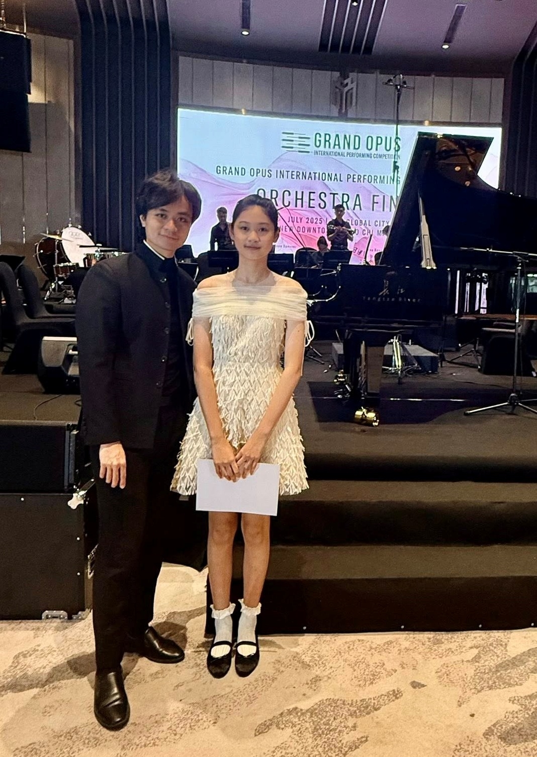 Tiểu Panda bên nhạc trưởng Dustin Tiêu tại buổi trình diễn thuộc cuộc thi Grand Opus Performing Competition - GOPC hồi tháng 7/2025. Anh nhận xét: Panda học piano không chuyên nhưng có tiếng đàn đẹp. Bé biết thả hồn vào tác phẩm, khả năng cảm nhạc tốt.