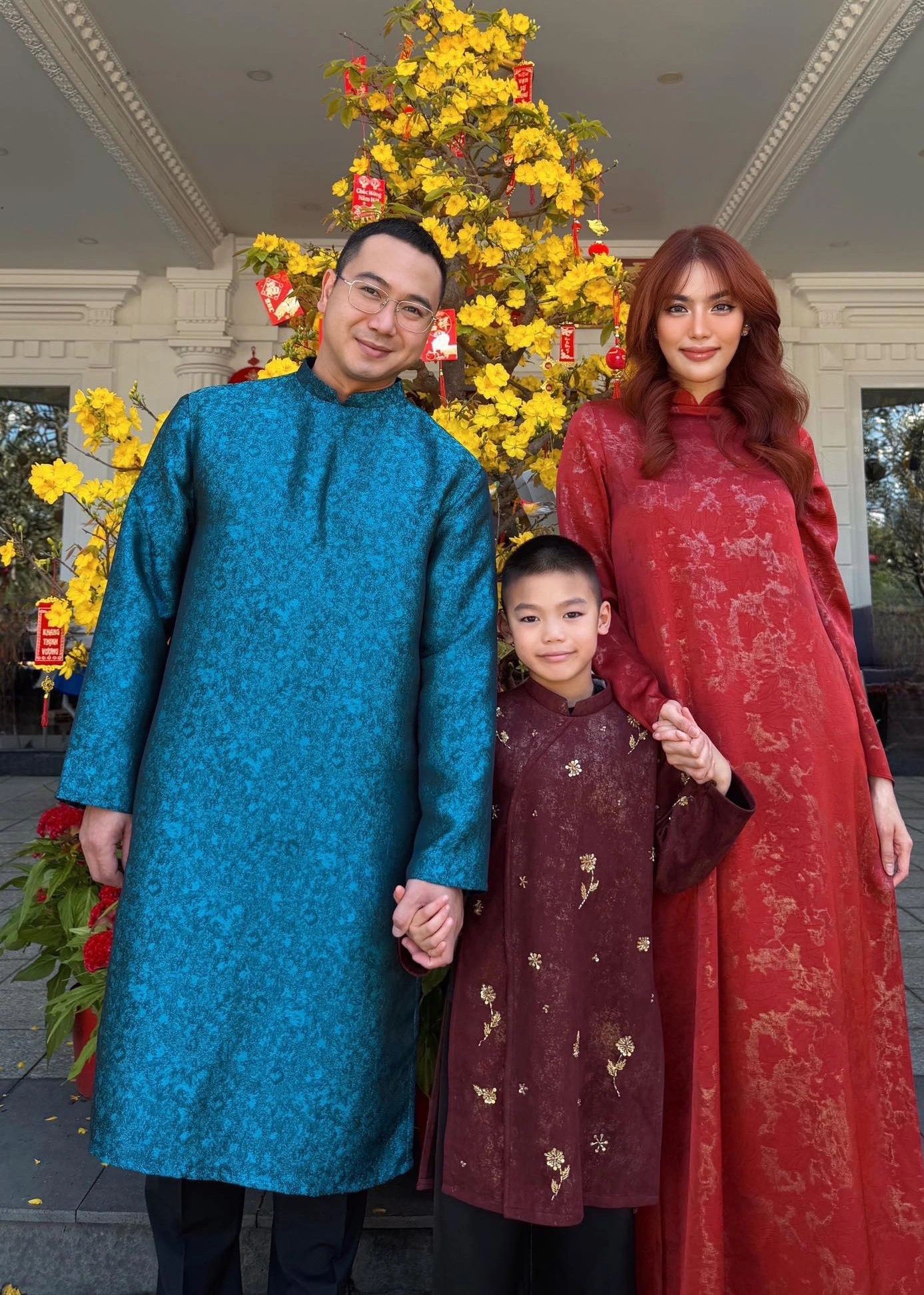 Dâu hào môn Lan Khuê xúng xính cùng chồng con trước cổng biệt thự. Cô lựa chọn áo dài đỏ ánh gấm, họa tiết vàng nhẹ tạo hiệu ứng thị giác bắt sáng. Mái tóc xoăn sóng bồng bềnh nhuộm đỏ rực tôn lên đường nét gương mặt nổi bật.