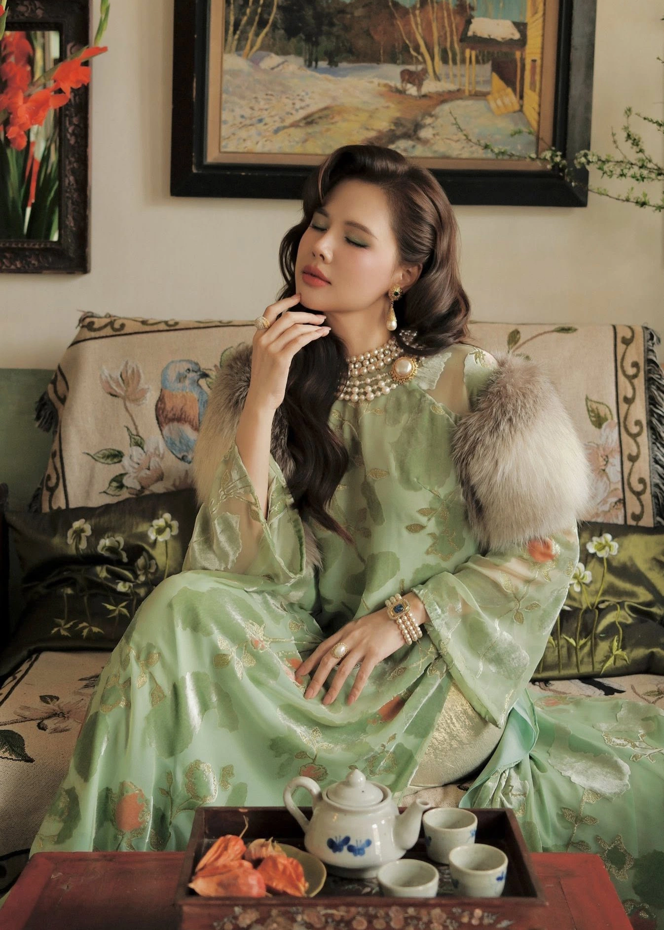Trong không gian nội thất đậm chất hoài niệm, Phanh Lee diện áo dài lụa xanh olive ánh kim phối cùng áo choàng lông sang trọng. Điểm nhấn nằm ở bộ trang sức ngọc trai đậm chất vintage, gợi nhắc phong cách quý phái của giới thượng lưu xưa. 