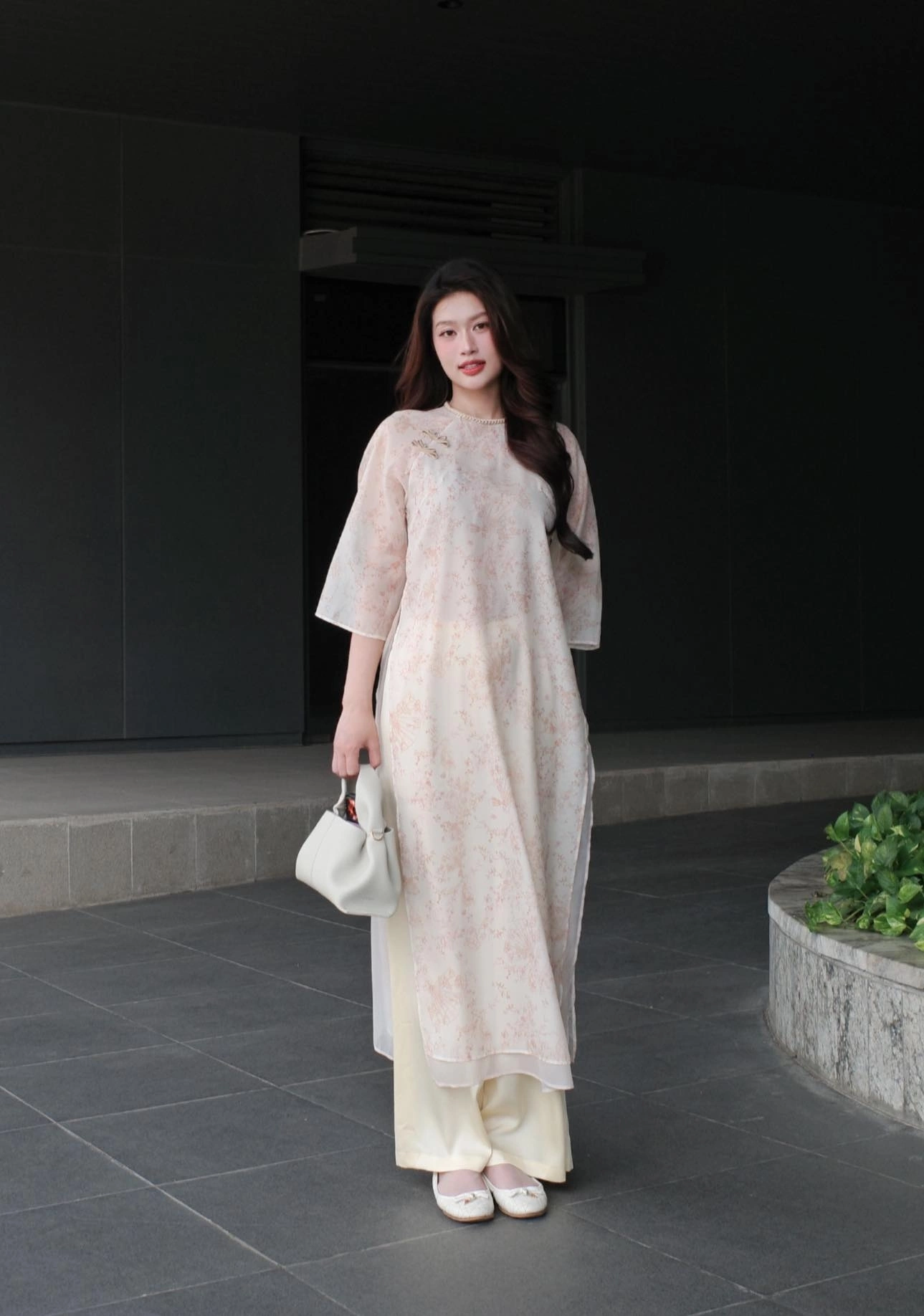 Hoa hậu Đoàn Thiên Ân duyên dáng trong mẫu áo dài lụa nền trắng sữa, điểm họa tiết hoa li ti. Thiết kế dáng suông mềm mại, tay lửng, kết hợp quần ống rộng cùng tông giúp tổng thể trở nên thanh thoát và trẻ trung. Đây là kiểu áo dài dành cho những cô gái yêu vẻ đẹp tối giản, tinh tế nhưng vẫn đủ cuốn hút để nổi bật giữa phố thị.