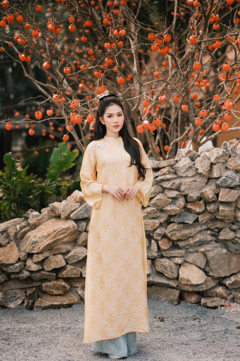 Mẫu áo dài vàng nhạt giúp tôn lên làn da trắng sáng của Ngọc Trinh. Họa tiết dệt chìm tinh xảo, tay loe mềm và phom dáng dài tạo cảm giác thướt tha, bay bổng theo từng chuyển động. Sắc vàng dịu không chỉ tôn da mà còn gợi liên tưởng đến sự may mắn, ấm áp và nữ tính, hợp không khí Tết.