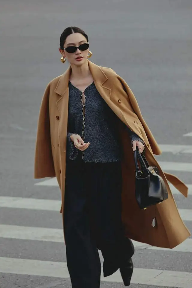 Áo khoác camel khoác hờ trên set đồ đen tạo nên tỷ lệ màu sắc hoàn hảo: ấm áp bên ngoài, tối giản bên trong. Nâu và đen là cặp đôi kinh điển, vừa quyền lực vừa thanh lịch. Phụ kiện vàng ánh kim nâng tầm tổng thể, mang lại cảm giác cao cấp mà không cần phô trương.