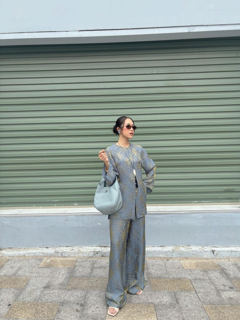 Một phiên bản áo bà ba hiện đại với dáng rộng, tay loe nhẹ, chất liệu gấm ánh kim màu xanh xám. Set đồ đồng bộ áo - quần tạo cảm giác phóng khoáng, thời thượng. Khi kết hợp cùng túi da tối giản, kính râm và trang sức bản nhỏ, áo bà ba bước ra khỏi khuôn khổ truyền thống để hòa mình vào nhịp sống đô thị.
