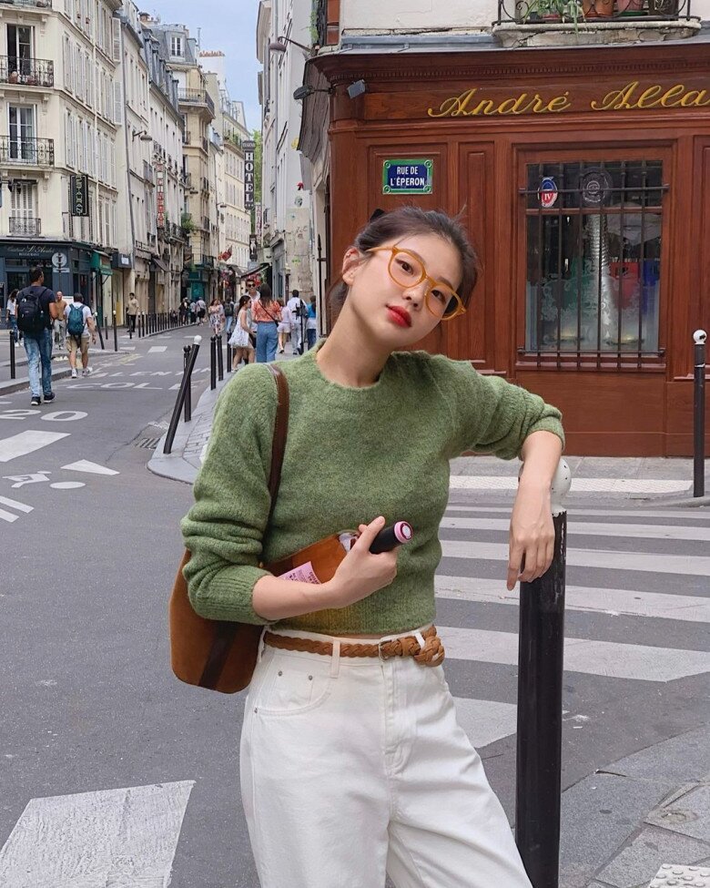 Áo len xanh lá giữ vai trò màu chủ đạo, tạo ấn tượng tươi mới. Quần trắng làm nền sáng, còn phụ kiện nâu như thắt lưng và túi xách đóng vai trò điểm nhấn ấm áp.