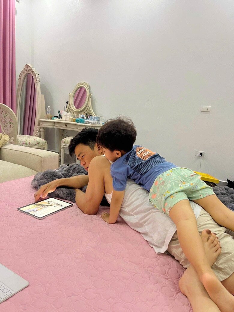 Phút giây thư giãn đời thường của hai cha con trên chiếc giường êm ái với gam hồng ngọt ngào. Phía xa là bàn trang điểm mang phong cách hoàng gia, phản ánh gu thẩm mỹ tinh tế của chủ nhân.