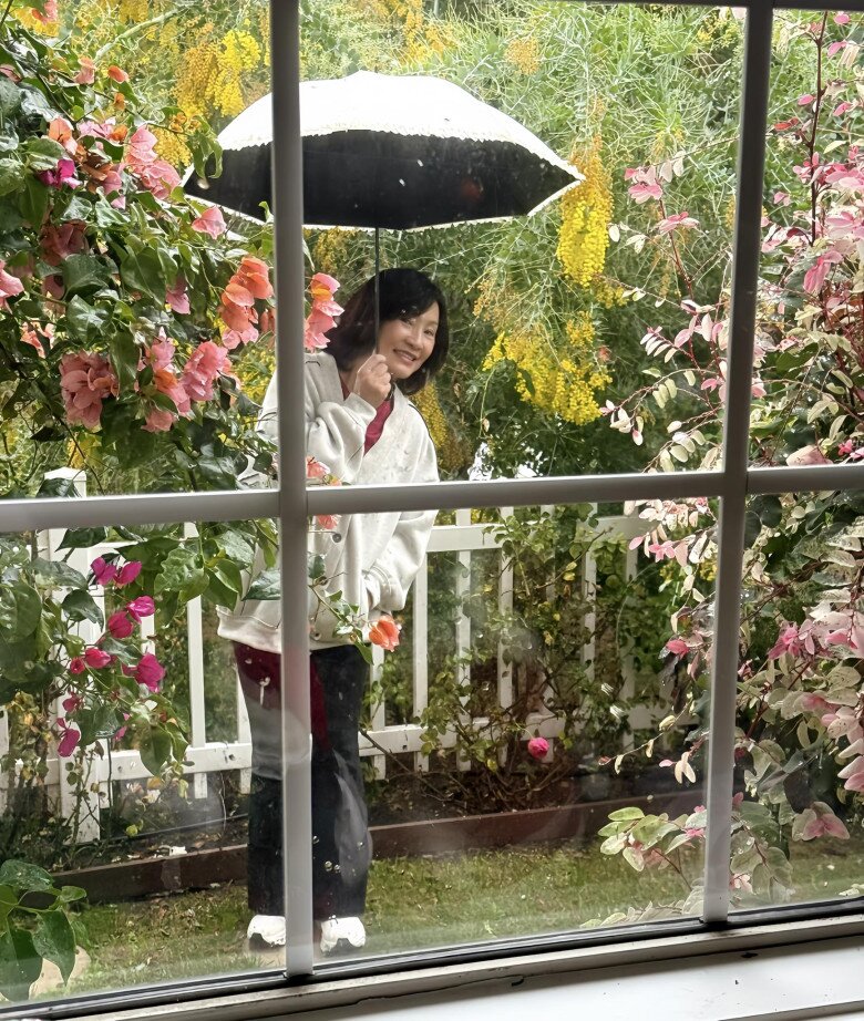 Hồng Đào đứng bên ngoài khung cửa, che ô dưới cơn mưa. Góc vườn rực rỡ sắc màu với những rặng hoa mimosa đan xen cùng sắc hồng đỏ của hoa giấy và cây xanh mơn mởn.