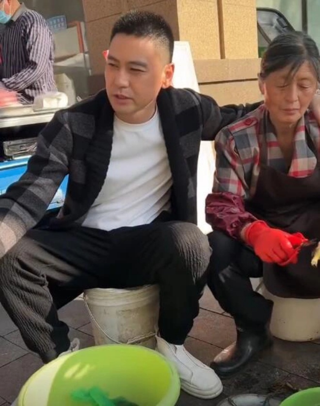Paradoxical male star in a suit sitting next to his mother in worn-out clothes selling fish in the market - 1
