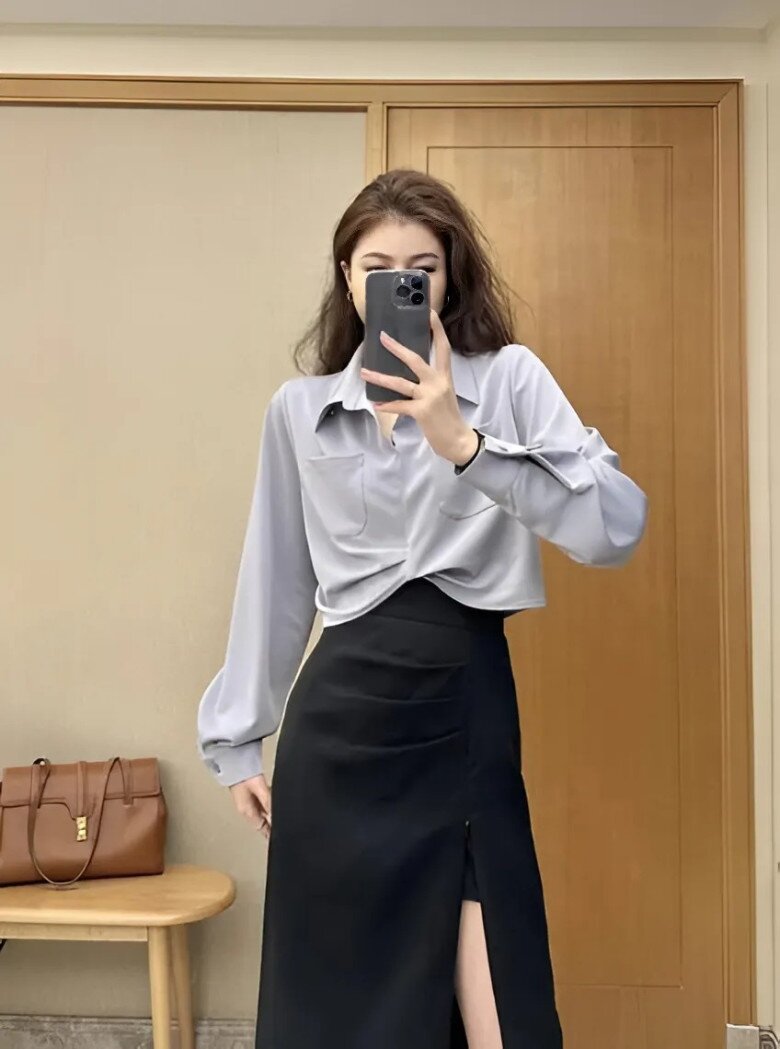 Chiếc sơ mi lụa được biến tấu bằng đường xoắn eo nhẹ nhàng, kết hợp chân váy đen xẻ cao, tạo nên tổng thể thời thượng và quyến rũ.