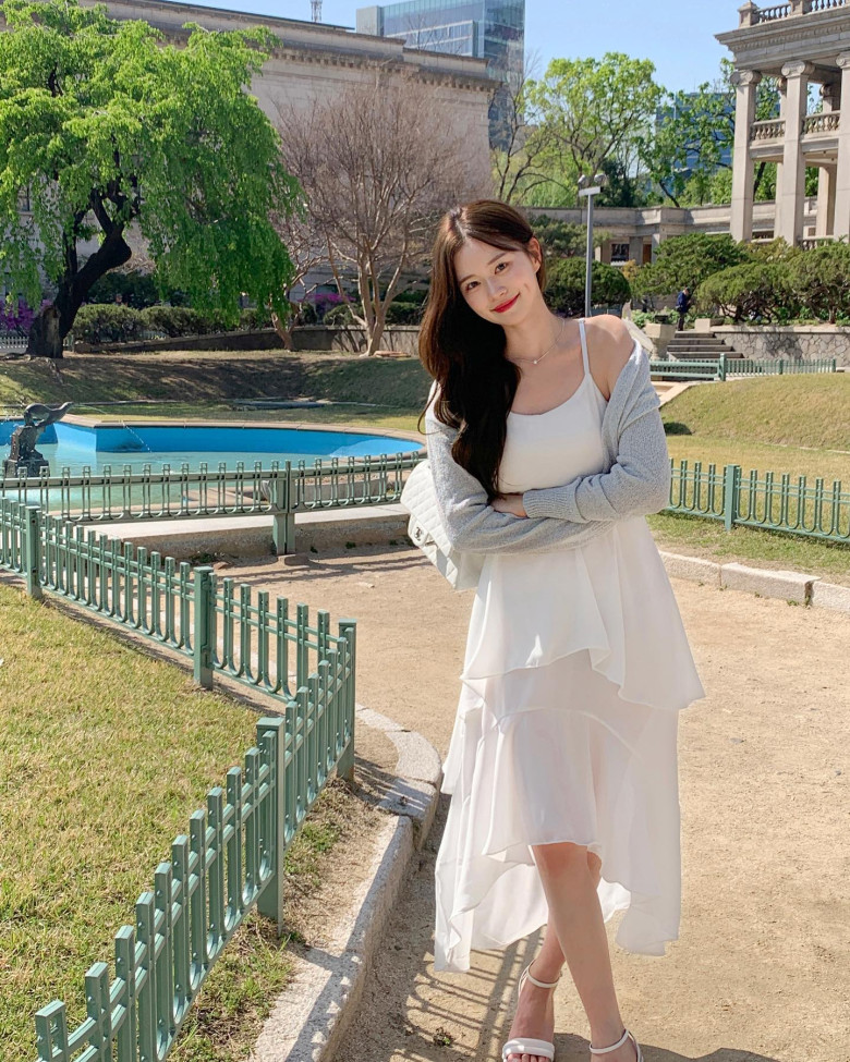 Nàng thơ dạo bước giữa nắng xuân với đầm trắng hai dây bay bổng, phối cùng áo len khoác hờ và sandals cùng tông giúp tổng thể thêm phần tinh tế, trẻ trung và trong trẻo như khung cảnh phía sau.