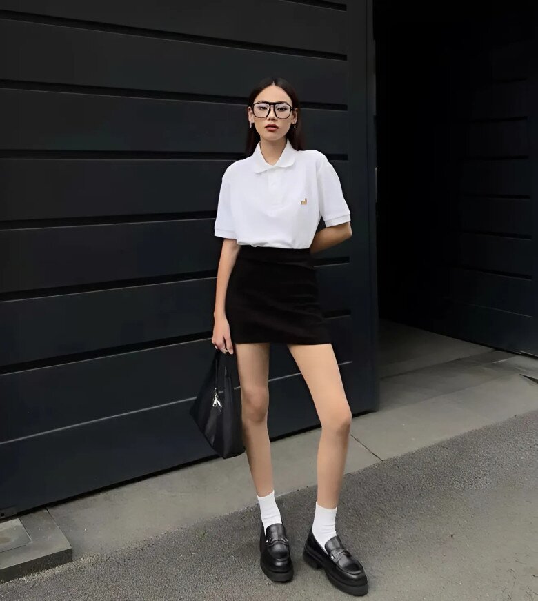 Áo polo trắng form rộng đi cùng chân váy đen ngắn ôm sát, phối với tất trắng và giày loafer tạo tổng thể cá tính.