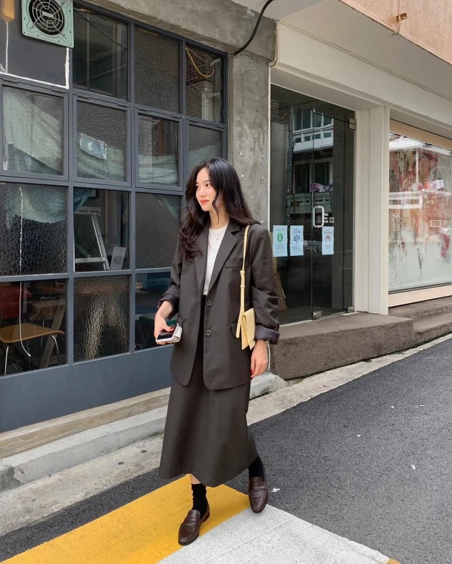 Chiếc váy dáng ôm dài tông trầm kết hợp cùng blazer oversized tạo nên tổng thể chuẩn “office chic”. Cách layering này gợi cảm hứng từ phong cách menswear, nhưng vẫn giữ nét mềm mại, thời thượng