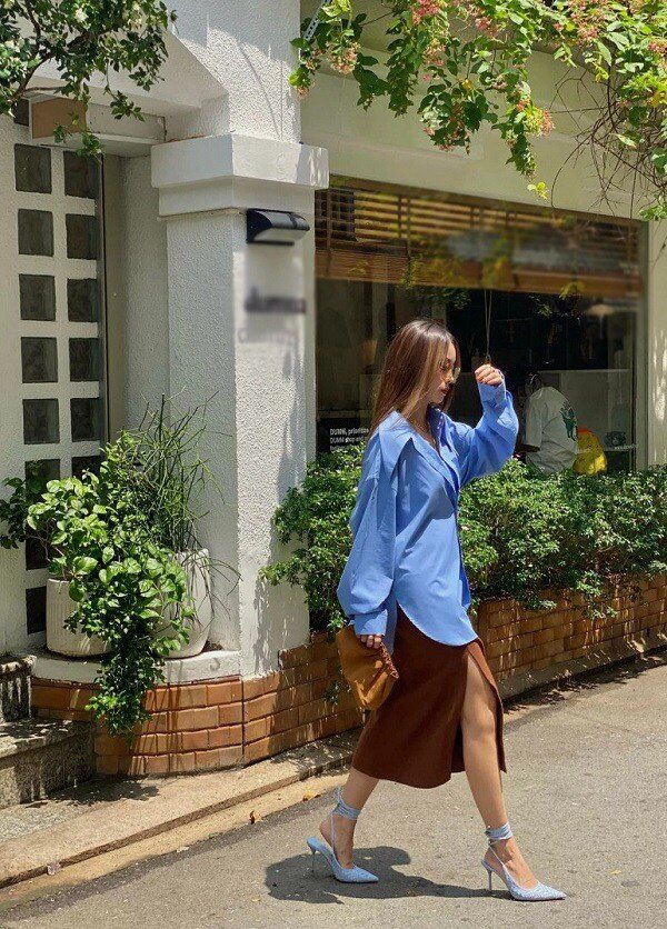 Sự kết hợp giữa sơ mi oversized xanh dương và chân váy lụa satin xẻ tà màu nâu mang đến nét chạm đầy phóng khoáng và thanh lịch. Điểm nhấn là đôi giày cao gót buộc dây ton-sur-ton với áo, tạo tổng thể hiện đại, gợi cảm nhưng không phô trương.