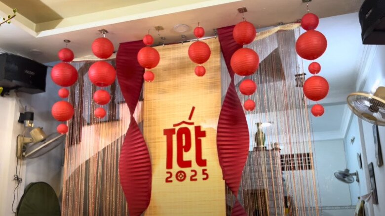 Phía sau là phông nền Tết với đèn lồng đỏ treo dọc lối đi được con gái của ông khéo tay trang trí. Màu sắc đỏ - vàng truyền thống gợi không khí lễ hội.