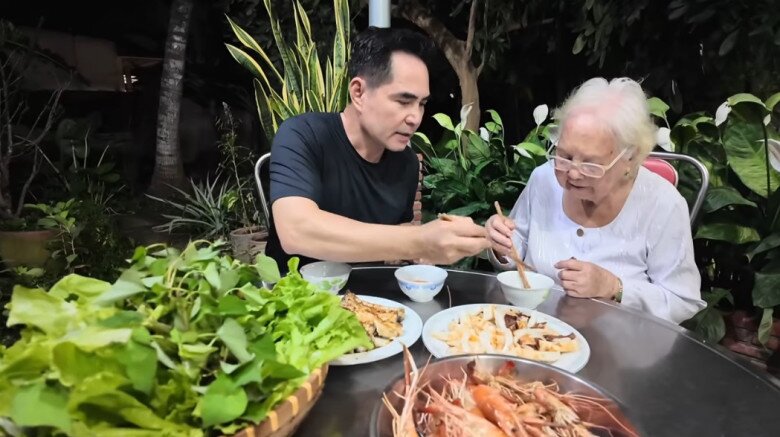 Khoảnh khắc ấm áp khi Trung Dũng ân cần gắp thức ăn mời mẹ. Hai mẹ con quây quần bên bữa ăn giữa sân nhà với rau xanh, cá nướng, mực tươi, tôm nướng – trọn vẹn hương vị quê nhà.