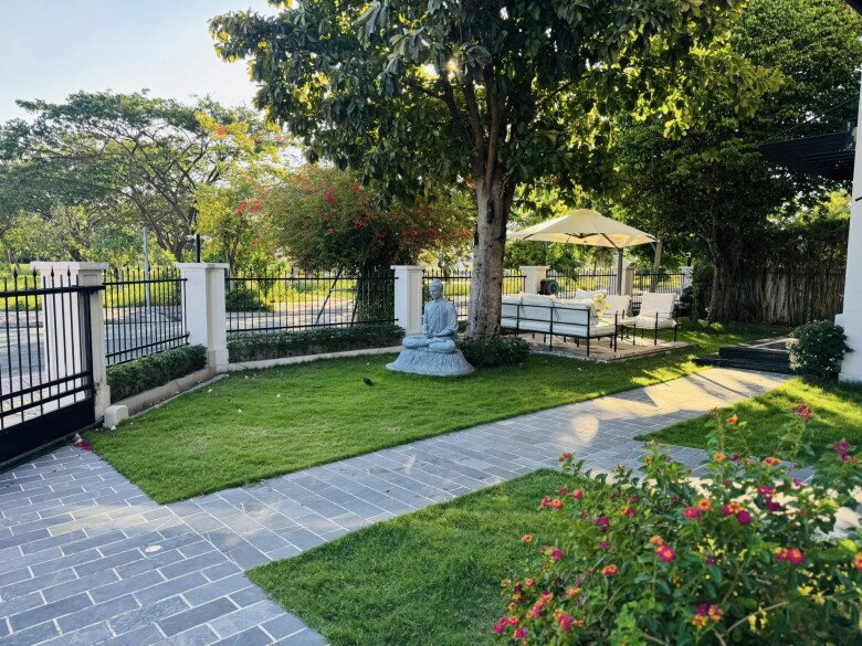 Bên trong biệt thự là khu sân vườn xanh mát, có tượng Phật đặt dưới gốc cây, phía xa là bộ bàn ghế trắng dưới dù che nắng – góc chill lý tưởng mỗi chiều hè.