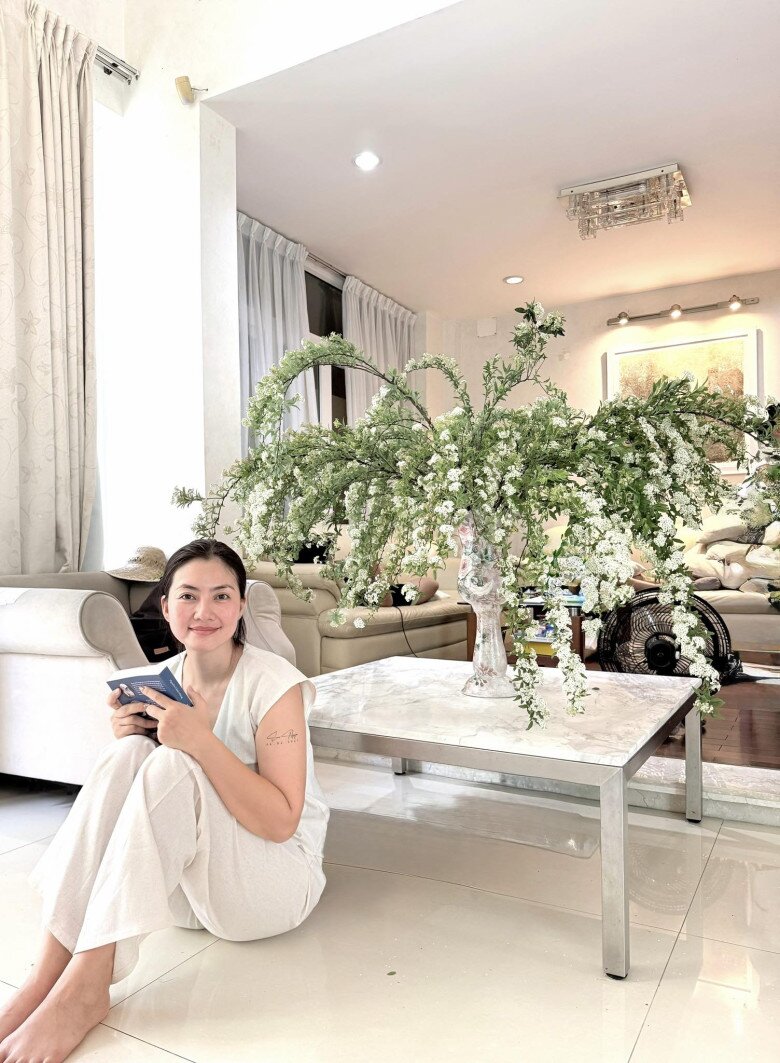Ngọc Lan ngồi thư giãn bên bình hoa lớn giữa phòng khách. Bộ sofa màu kem, rèm trắng và ánh sáng tự nhiên tạo nên khung cảnh sáng sủa, yên bình – đúng tinh thần “nhà là nơi để thở”.