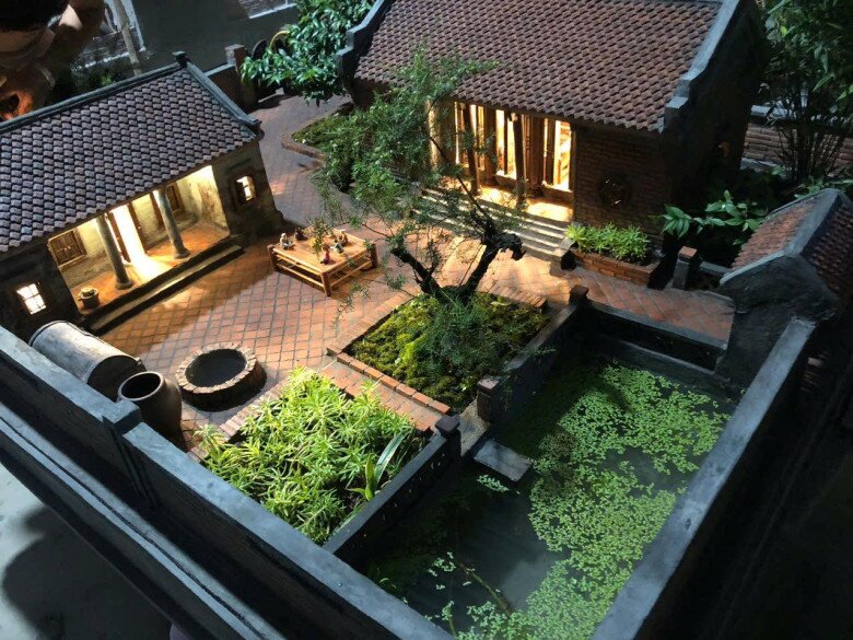 Cận cảnh mô hình với sân lát gạch đỏ, giếng nước, cây cảnh và ao nhỏ, tạo nên một tổng thể bình dị, gần gũi. Ánh sáng từ bên trong ngôi nhà khiến không gian thêm ấm áp.