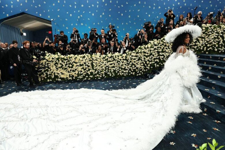 Diana Ross tái xuất ở Met Gala sau hơn 20 năm với thiết kế đầm lông vũ, kèm áo choàng thêu tên của tất cả các con và cháu của cô. Vì bộ váy quá sức đồ sộ nên cần phải có 4 người trợ lý giúp cô nâng áo lên bậc thang.