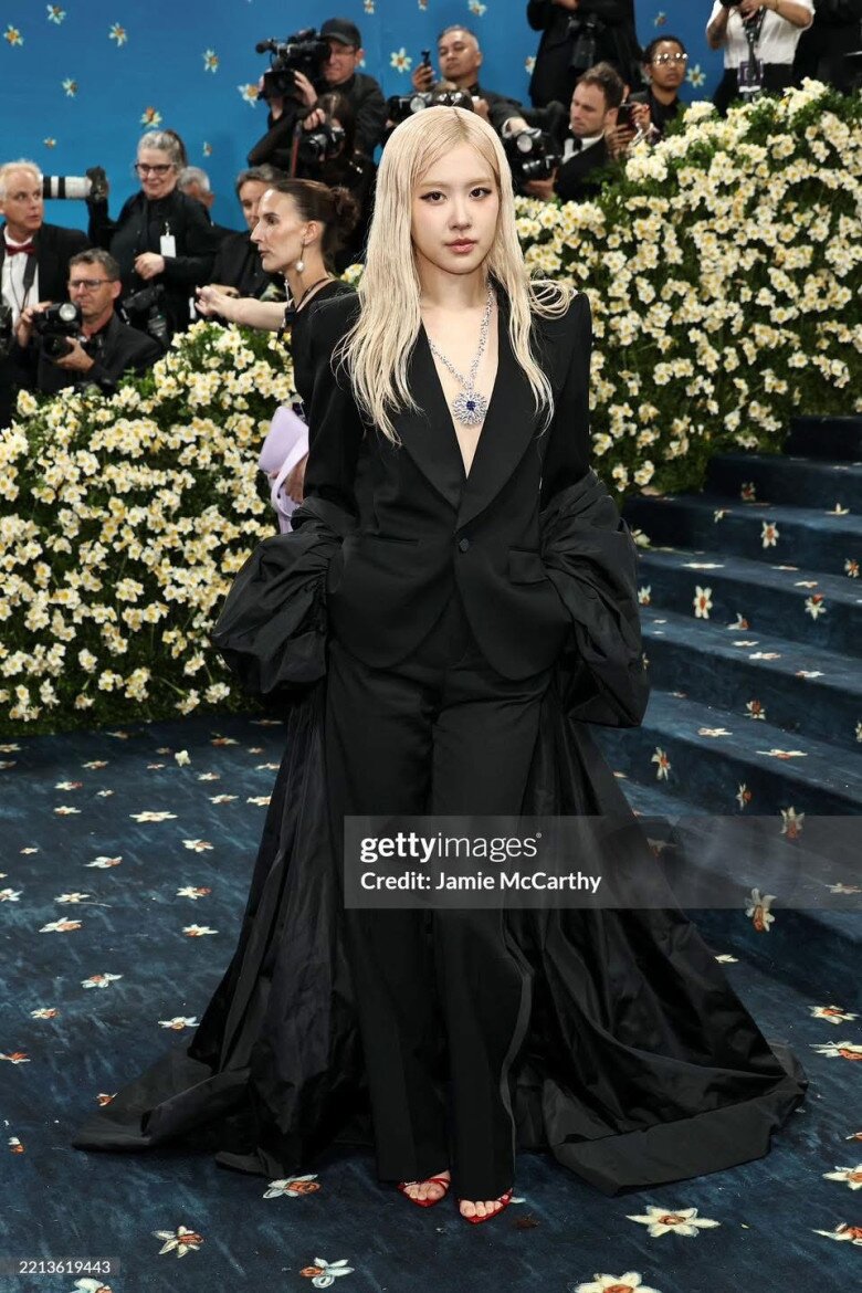 Rosé diện một bộ suit đen hoàn toàn của Saint Laurent. Bộ suit được may đo sắc nét, với áo vest cổ V sâu, để lộ phần ngực tinh tế, kết hợp cùng quần ống loe dài, tạo phom dáng mạnh mẽ nhưng vẫn nữ tính. Điểm nhấn là chiếc áo choàng đen dài, làm từ vải satin bóng mượt, quét đất.