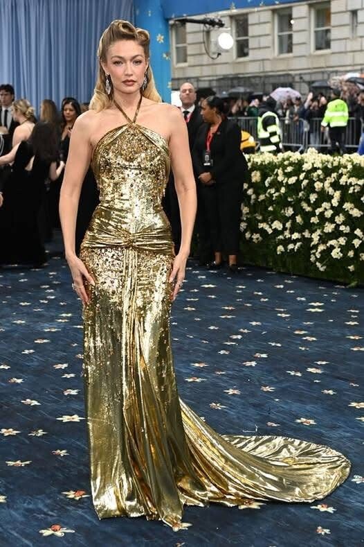Gigi Hadid diện đầm metallic, dù chẳng hở hang nhưng mỹ nhân 9X vẫn phô diễn được body không mỡ thừa. Gigi chọn phong cách trang điểm gợi nhớ đến minh tinh Marilyn Monroe, với đôi môi đỏ rực rỡ và má hồng nhẹ, tạo vẻ quyến rũ cổ điển. Đôi mắt được kẻ eyeliner mèo mảnh, nhấn nhá bằng phấn mắt ánh kim nhẹ, làm nổi bật ánh nhìn.