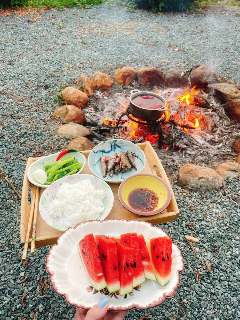 Trải nghiệm ẩm thực dân dã và ấm cúng của gia đình giữa không gian thiên nhiên hoang sơ. Mâm cơm đơn sơ với cá kho, rau luộc, trứng và dưa hấu được bày biện khéo léo trên khay gỗ, thưởng thức ngay cạnh bếp củi bập bùng giữa sân sỏi, mang lại cảm giác nguyên sơ, thư thái.