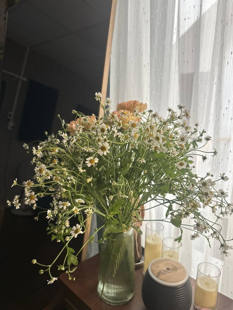 Góc làm việc bừng sáng với bình hoa cẩm chướng hồng phấn dịu dàng đan xen cùng những đóa cúc tana trắng muốt, tạo cảm giác thư thái và khơi nguồn cảm hứng sáng tạo cho nữ ca sĩ. Ánh sáng tự nhiên từ cửa sổ hắt vào làm tôn lên vẻ đẹp mong manh nhưng đầy sức sống của loài hoa đặc trưng của mùa thu.