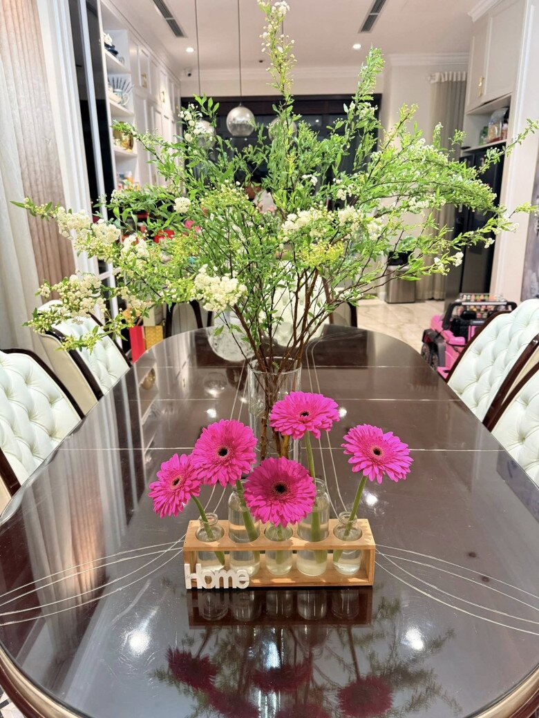 Những bông đồng tiền màu hồng đậm được cắm riêng biệt trong lọ nhỏ dạng ống đặt trên giá gỗ mộc mạc. Phía sau là những cành hoa thanh liễu trắng li ti tạo nên lớp nền bồng bềnh cho bàn ăn.