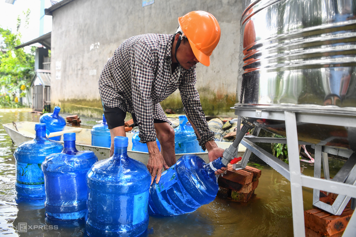 Ông Nguyễn Văn Tho chèo thuyền từ thôn Đồng Rạch ra điểm lấy nước cách nhà hơn một km. Ông mang theo 10 bình loại 20 lít để lấy nước phục vụ sinh hoạt cho gia đình 5 người. Chỗ tôi ngập hơn 2 m, hôm qua lấy 4 bình nhưng không đủ dùng nên nay phải lấy thêm, ông Tho nói, xòa đôi bàn tay nhăn nheo vì ngâm nước.