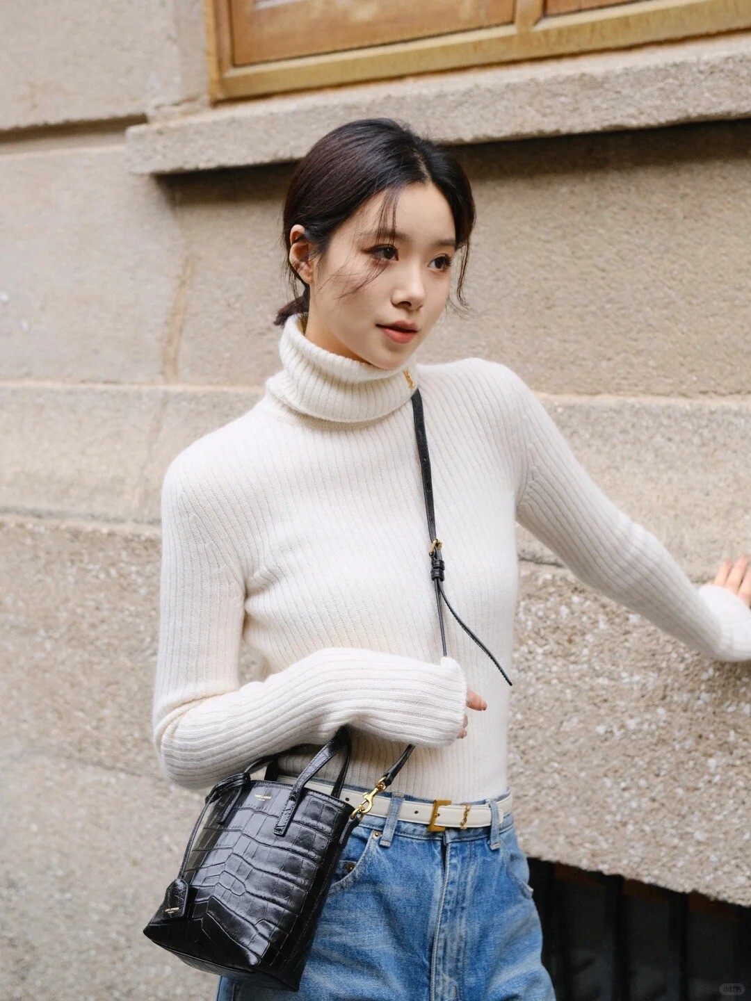 A woman wearing a white turtleneck sweater and blue jeans, standing in front of a colorful mural.