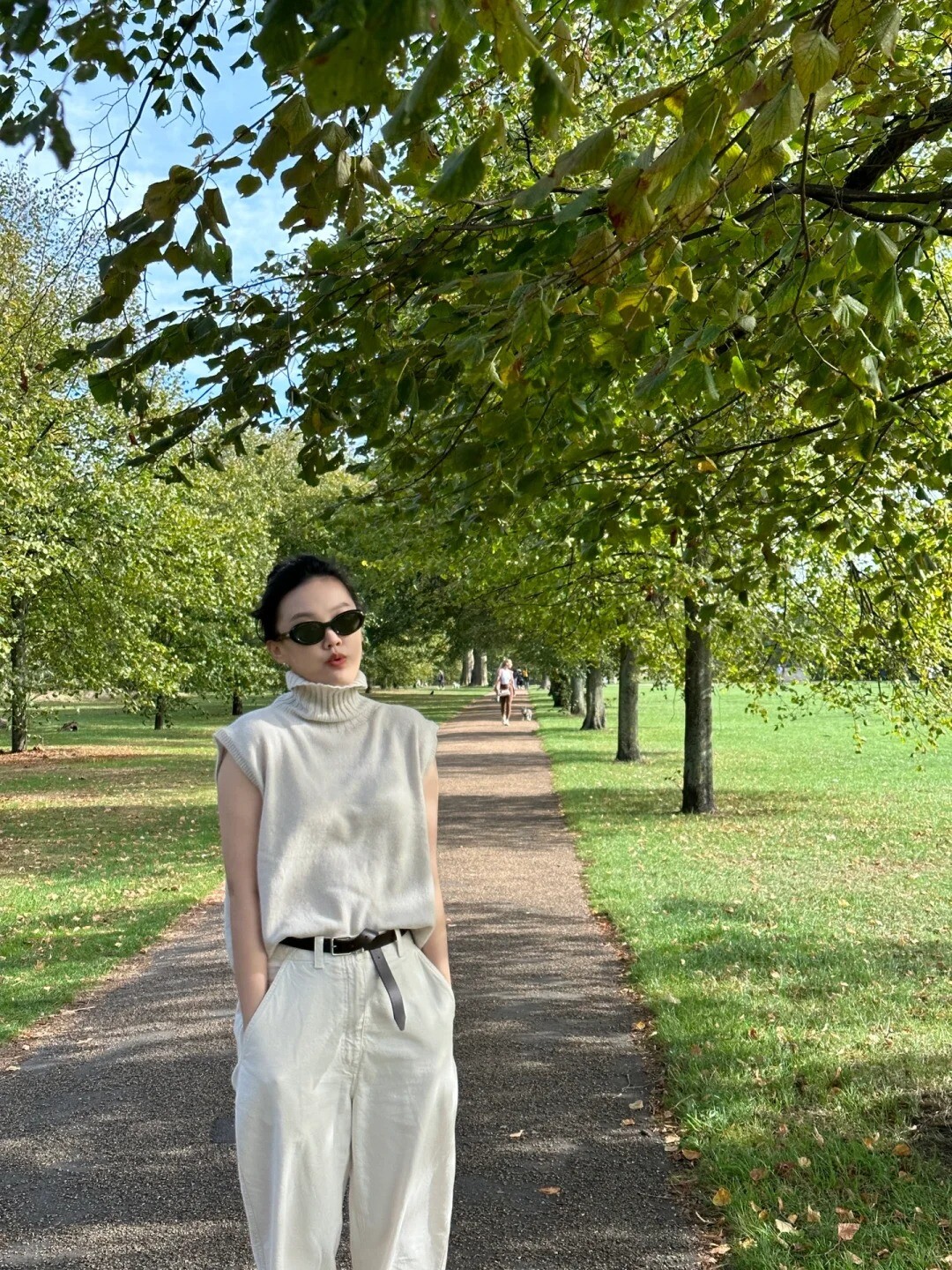 A woman wearing a black sleeveless sweater, white shirt, black pants, and white sneakers, standing in a cityscape.