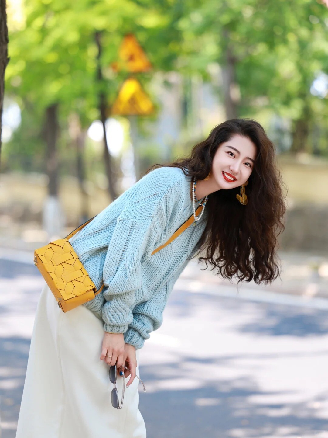 A woman wearing a bright yellow sweater, blue jeans, and white sneakers, posing on a street.