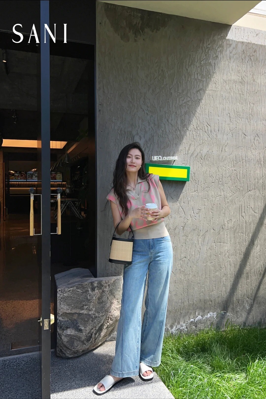 A woman wearing a beige sleeveless sweater, white shirt, and blue jeans, posing on a street.