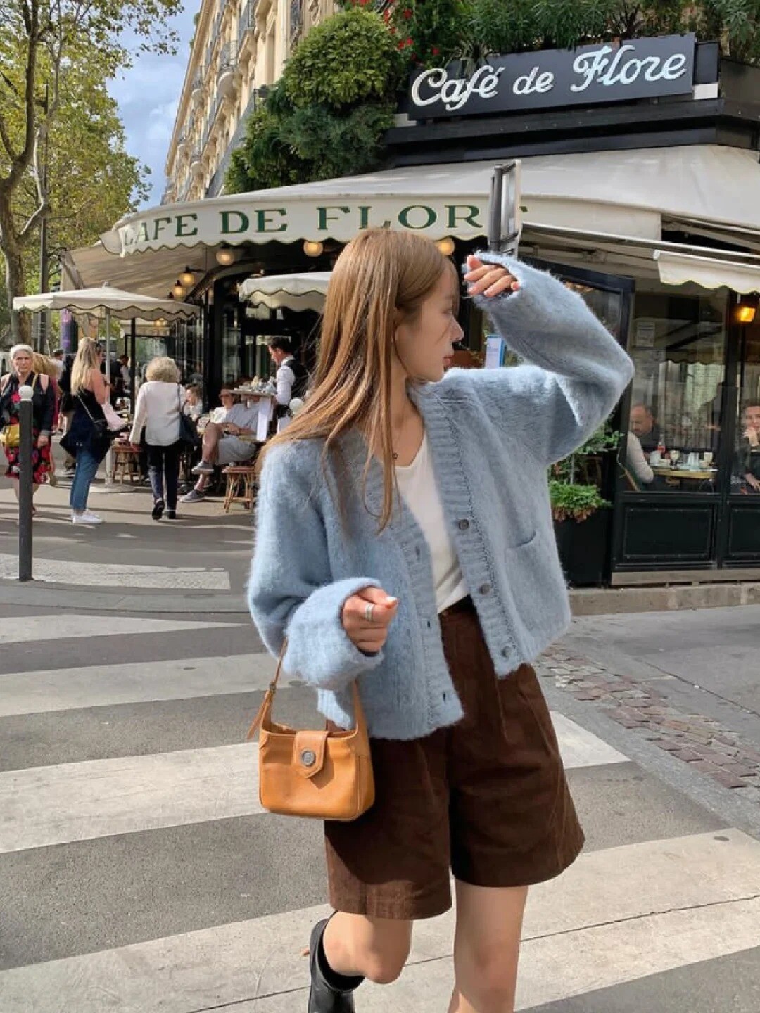 A woman wearing a beige cardigan, white top, and blue jeans, posing on a street.