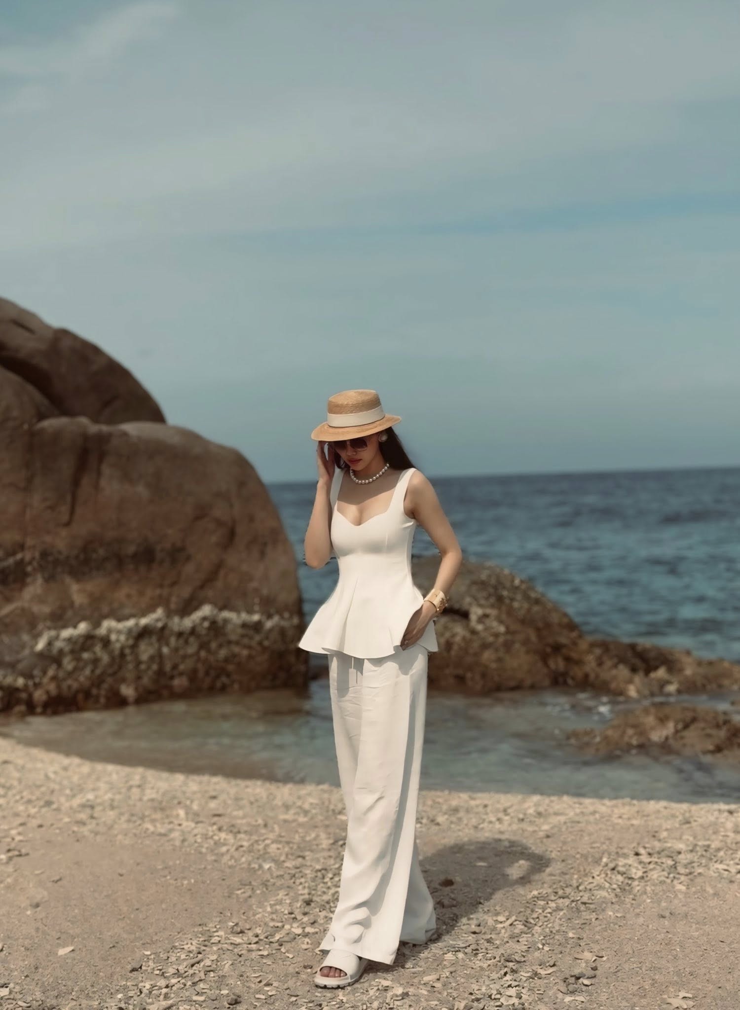 Điểm nhấn của set đồ nằm ở cách phối phụ kiện đầy tinh tế. Mũ cói boater mang hơi thở cổ điển, kết hợp cùng chuỗi vòng cổ và khuyên tai ngọc trai tạo cảm giác sang trọng nhưng vẫn rất phù hợp với khung cảnh biển trời. Cô hoàn thiện outfit bằng sandal trắng và vòng tay bản lớn màu vàng ánh kim của Chanel có giá hơn 25 triệu, vừa đủ nổi bật nhưng không hề phô trương.