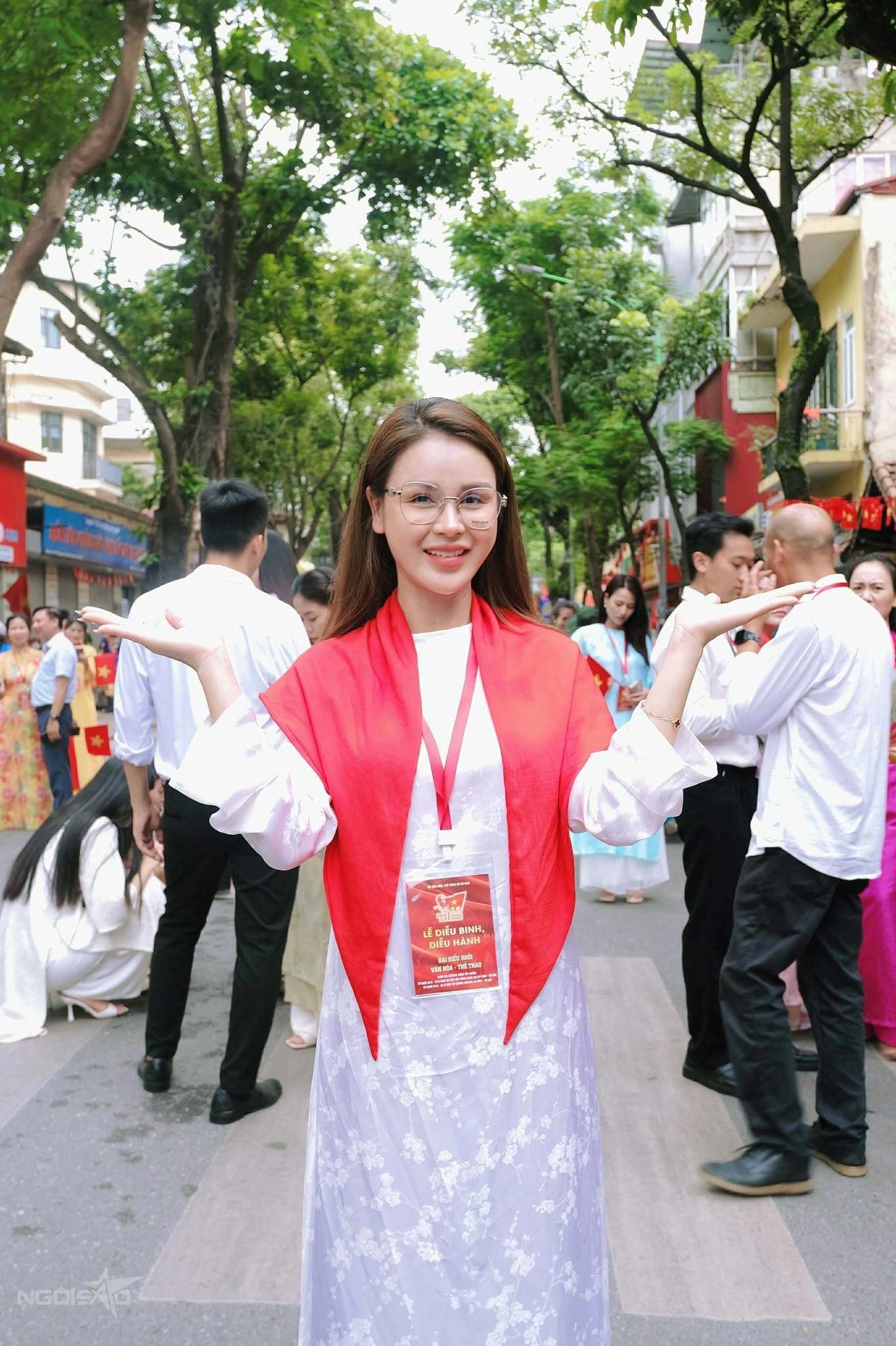 Nữ diễn viên Lương Thu Trang diện áo dài trắng họa tiết hoa nhẹ, kết hợp khăn choàng đỏ tạo điểm nhấn nổi bật. Cặp kính trong suốt mang đến vẻ hiện đại xen lẫn truyền thống, giúp tổng thể vừa trẻ trung, vừa duyên dáng khi xuất hiện trong không khí lễ hội rộn ràng.