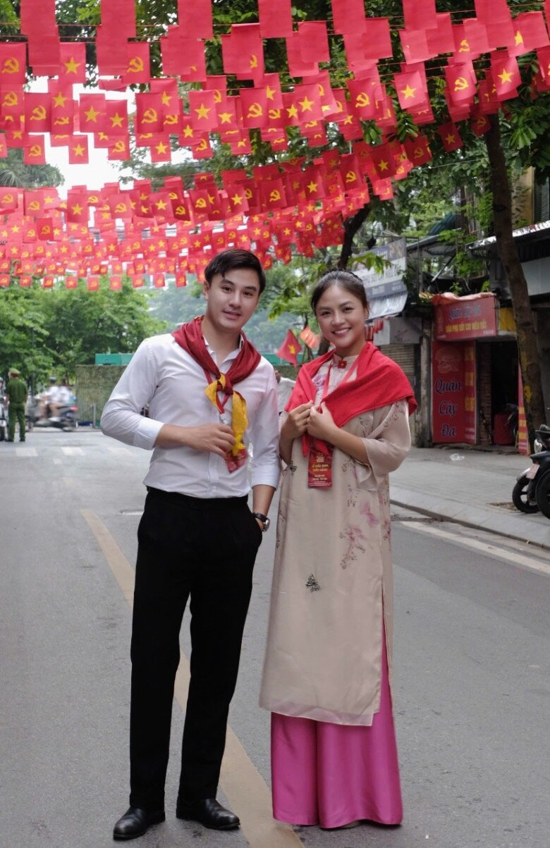 Thu Quỳnh chọn áo dài gấm be in hoa nhẹ, phối quần lụa hồng nổi bật. Điểm nhấn khăn choàng đỏ đồng điệu với bối cảnh rợp cờ, tạo nên hình ảnh vừa truyền thống, vừa trang nhã.