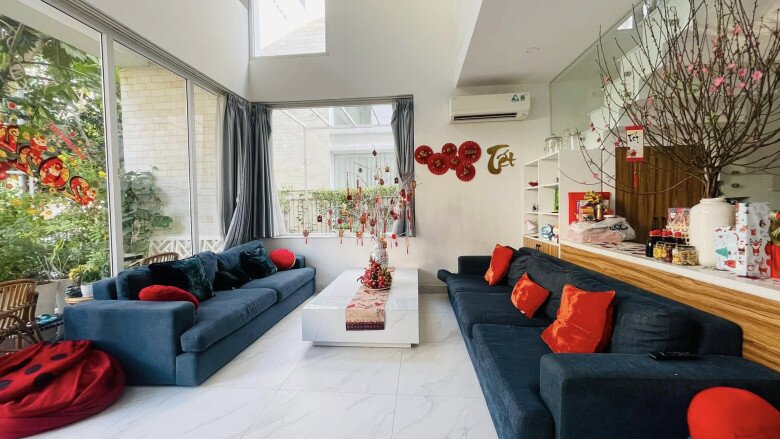 Hai dãy ghế bố trí đối xứng quanh bàn trà mặt kính trắng, trên bàn là mảng trang trí đơn giản nhưng gọn gàng. Tường kính giúp ánh sáng và mảng xanh từ ngoài vườn ùa vào phòng, mang thiên nhiên sát bên chỗ ngồi.