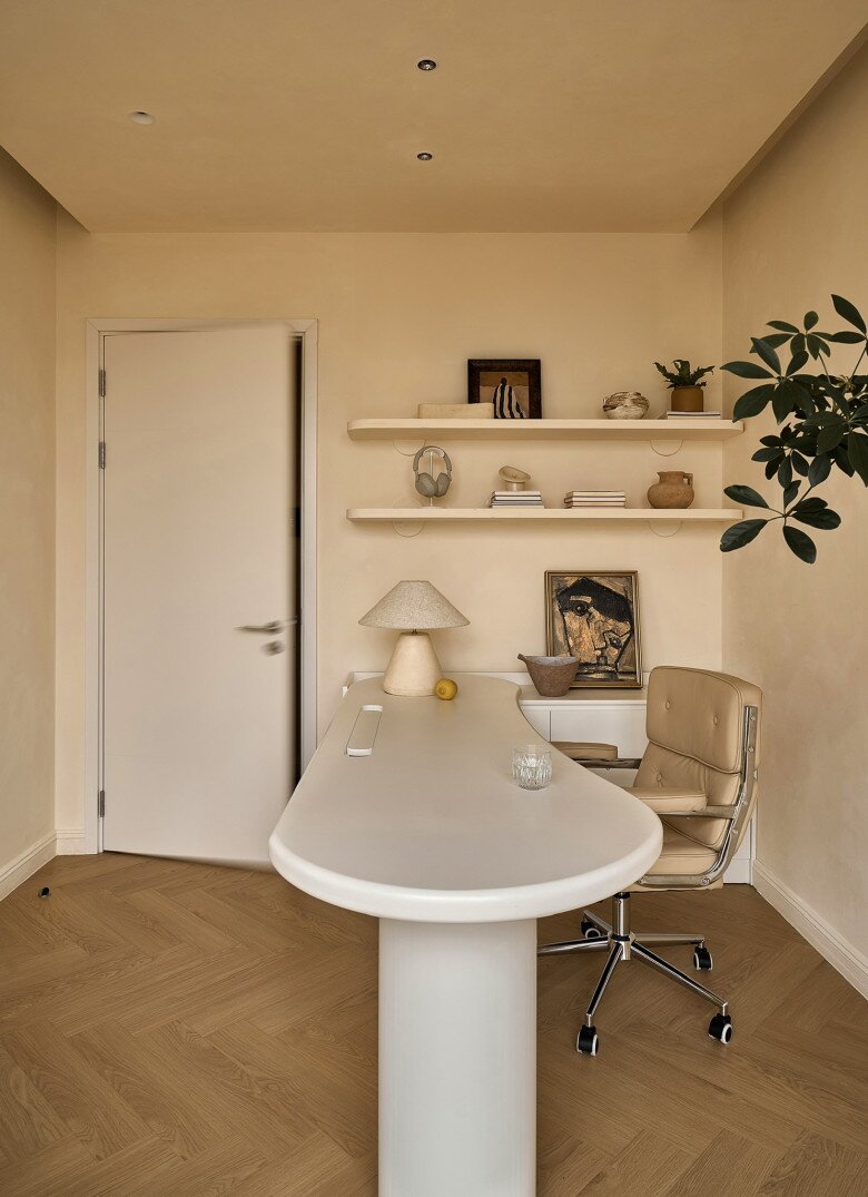 Hai tầng kệ nổi treo tường trưng bày sách và vật phẩm trang trí. Chiếc ghế xoay da màu beige và đèn bàn gốm là những chi tiết làm tăng độ “mướt” cho không gian.