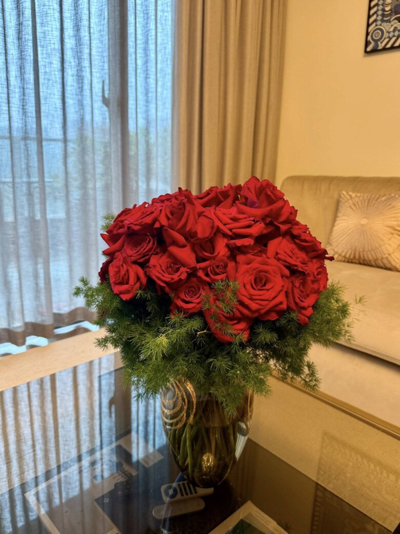 Bình hoa hồng đỏ rực được nam diễn viên cắm tròn đều, phối kèm lá kim xanh tạo nền tôn hoa. Tone đỏ nổi bật giữa không gian trung tính, đem lại cảm giác ấm cúng, sang trọng.