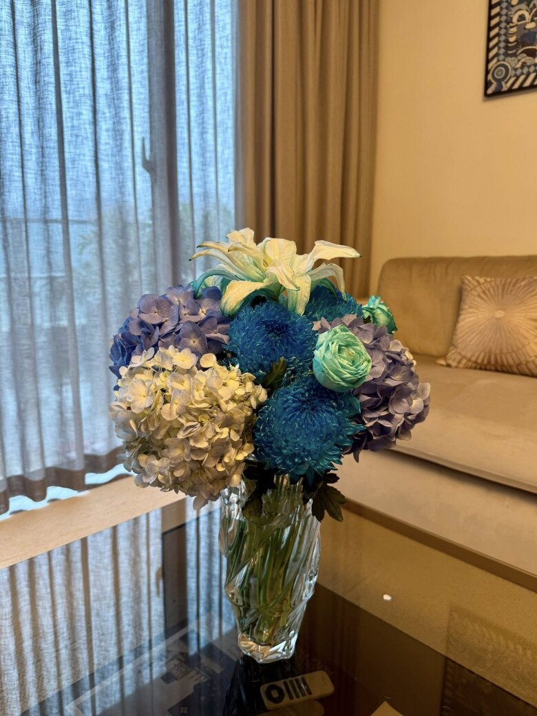 Hoa xanh – trắng – tím được anh cắm nhẹ nhàng trong bình pha lê, mang hơi hướng thanh mát, dịu dàng. Bố cục cân đối, tạo điểm nhấn hài hòa trong không gian phòng khách sáng tự nhiên.