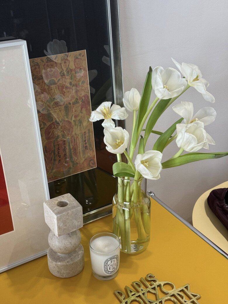 Hoa tulip trắng cắm trong lọ thủy tinh đơn giản, đặt trên mặt bàn vàng cùng nến thơm, decor theo phong cách Bắc Âu. Tối giản nhưng không hề đơn điệu.