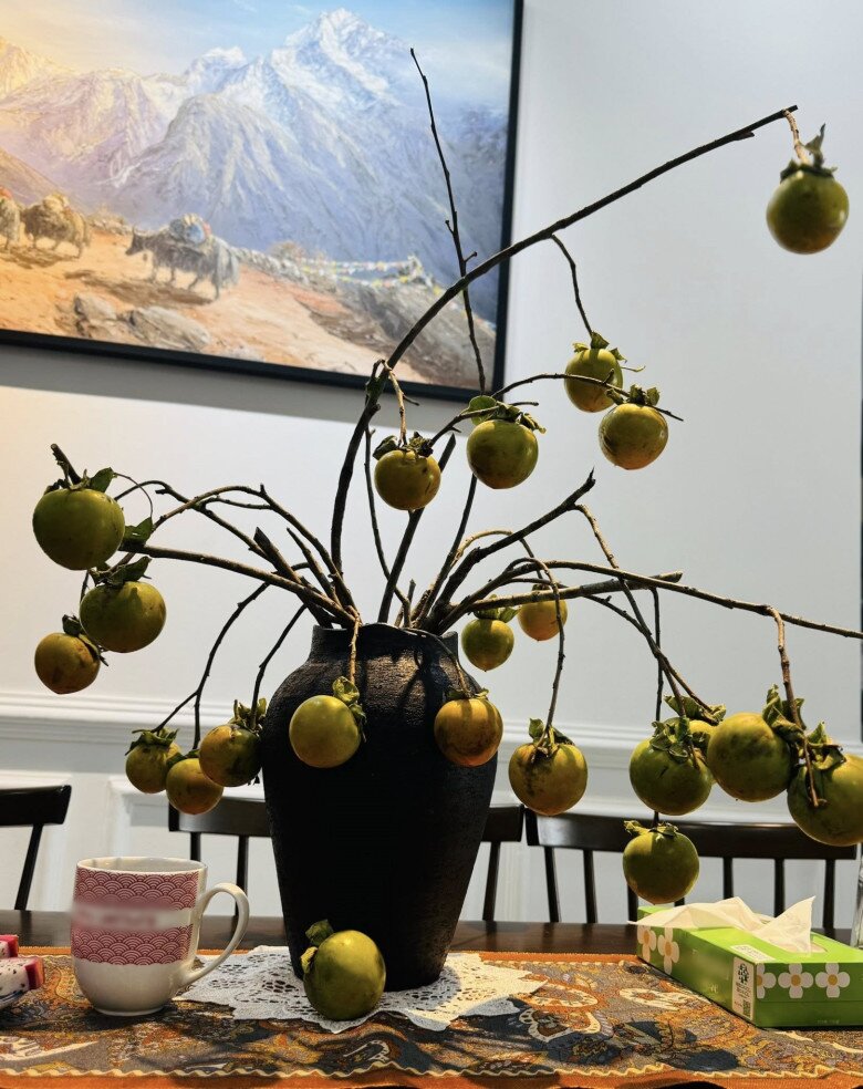 Góc bàn ăn với ghế gỗ và tranh treo tường đơn sắc được điểm xuyết bởi bình hoa hồng tròn tán, quả lớn và đều. Một tách trà và hộp khăn giấy được đặt bên cạnh, gợi cảm giác thư giãn, bình dị.