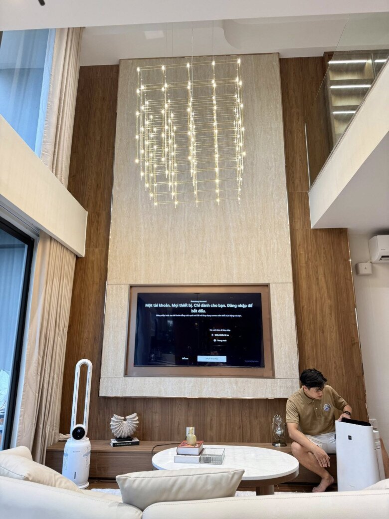 Tường gỗ kết hợp mảng đá sáng màu phía sau TV làm bật lên sự sang trọng. Đèn LED dạng lưới thả từ trần cao tới giữa phòng vừa hiện đại vừa tạo hiệu ứng thị giác độc đáo. Các món decor nhỏ như tượng nghệ thuật, nến và sách decor càng làm tăng sự chỉn chu cho không gian.